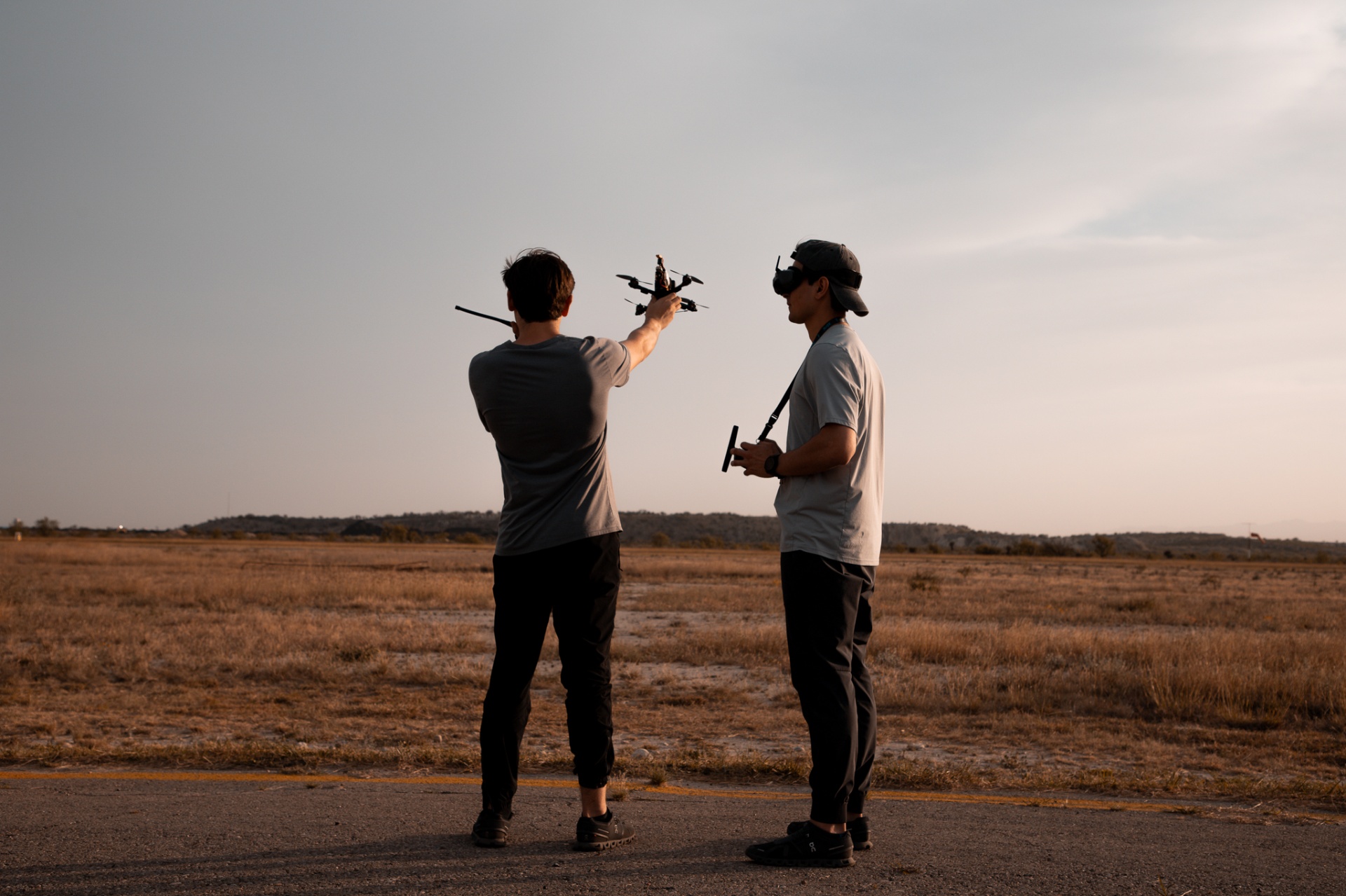 Two FPV operators hand-launching a cinelifter on a desert strip at golden hour