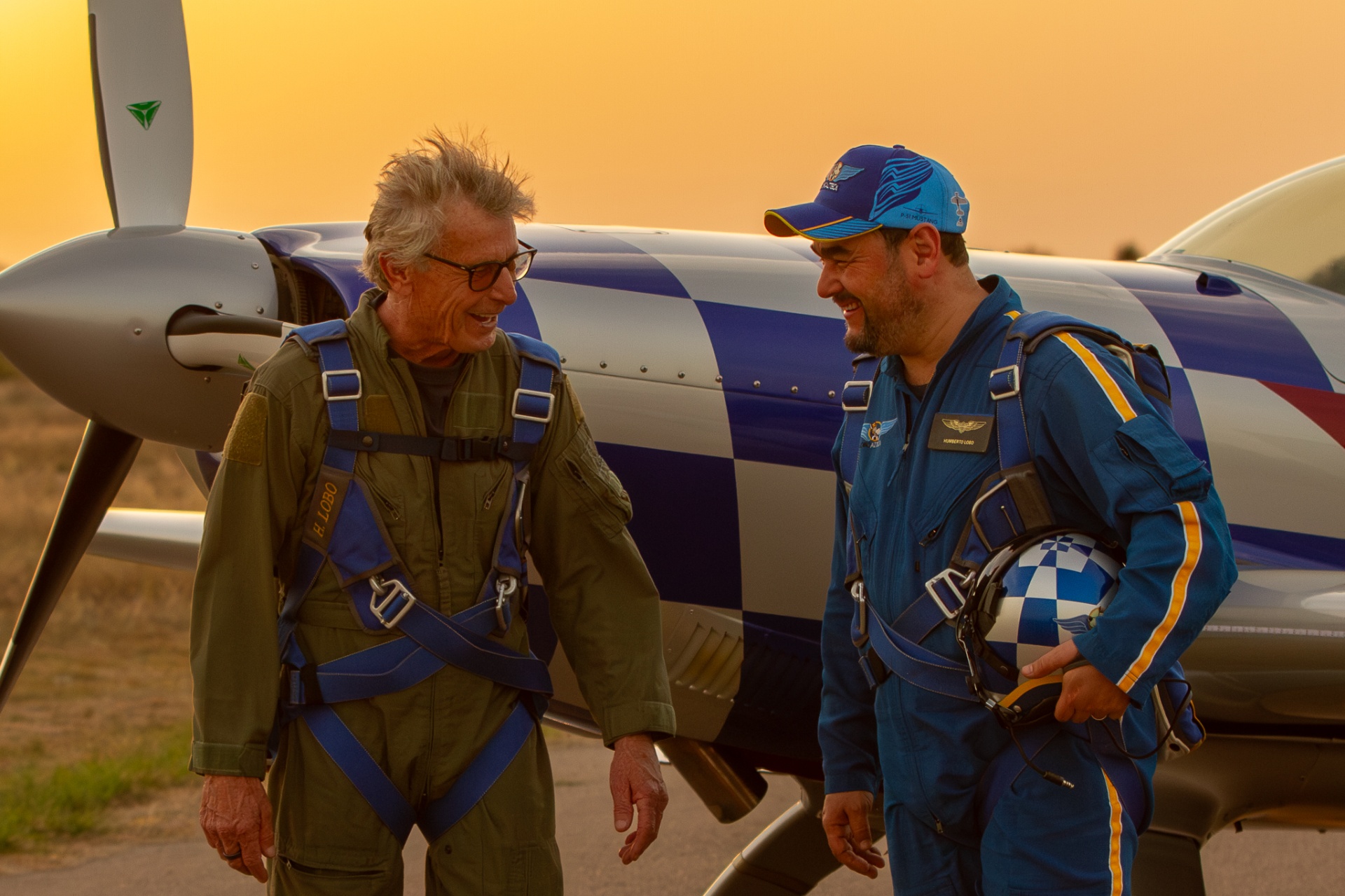 Two pilots in flight suits laughing in front of blue-and-silver Extra aerobatic plane